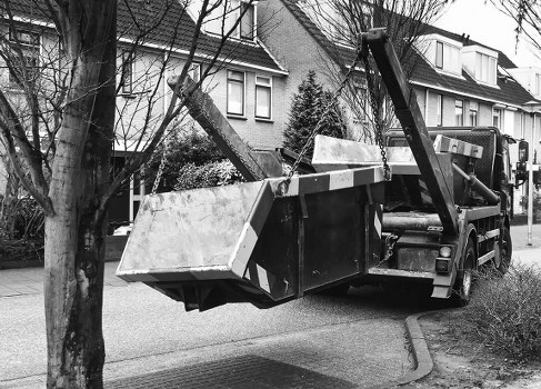 A flatbed tow truck with a hydraulic lifting arm is parked on a residential street, lifting a large, rectangular metal container filled with mixed rubbish and debris. The container appears weathered with visible dirt and corrosion, and is secured to the truck's flatbed which has a textured black surface. The street is lined with modern residential houses, some with front gardens and trees, and the pavement runs alongside. The background features leafless trees and rooftops of several houses, indicating an urban or suburban area. The scene is captured in black and white, emphasizing the industrial nature of the rubbish removal process, which is handled by Commercial Waste Removal Southgate, offering commercial rubbish clearance services in the local vicinity.