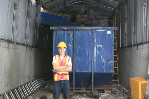 A worker wearing a yellow safety helmet and a high-visibility vest stands with arms crossed in front of a large, weathered blue metal skip filled with mixed waste materials inside an industrial waste storage area. The skip has a corrugated surface with visible scratches and signs of use, positioned on a concrete surface scattered with small debris and waste. To the right, there is a yellow plastic container, and to the left, a folded metal ladder leans against the wall. The interior of the container is sheltered by a semi-open roof structure made of metal and translucent panels, with grey metal siding on either side. Environmental details suggest a waste management facility or a commercial rubbish collection point, consistent with services offered by Commercial Waste Removal Southgate near London, addressing rubbish removal needs in the Southgate area.