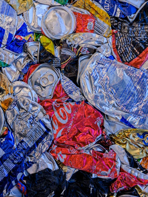 A close-up view of a pile of crushed and crumpled aluminum cans, primarily consisting of soft drink and soda cans with metallic silver surfaces, variously painted with red, blue, black, and yellow bra