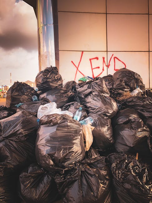 A black, cast-iron trash bin with a domed lid and vertical ridges stands on a cobblestone street, surrounded by scattered litter including glass bottles, paper, and plastic wrappers at its base. On to