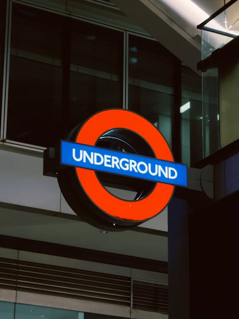 A view of the underground platform at a tube station showcasing the curved tiled ceiling with bright fluorescent lighting running along its center. On the platform, there are several seated and standi