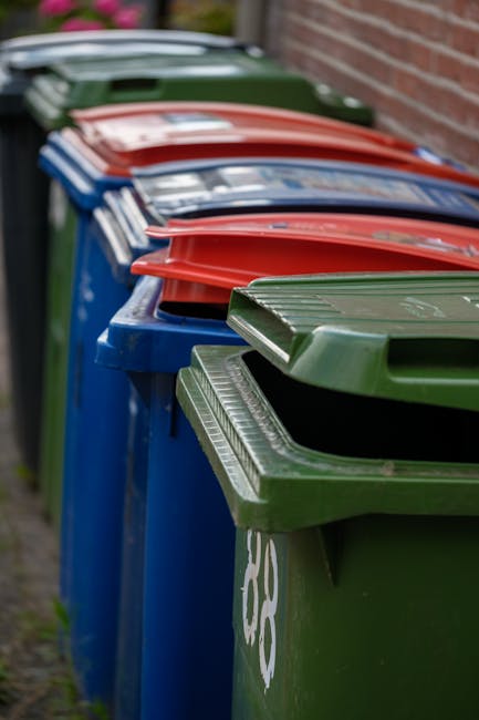 A large, weathered red metal rubbish skip with visible rust and scratches, placed against the corner of a building’s exterior wall. The skip is situated on a dark paving surface and is partially overf
