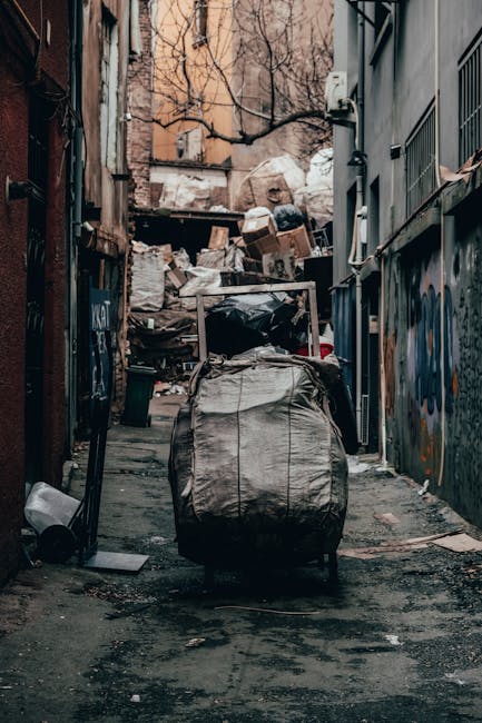 Two large black plastic garbage bags filled with waste materials are positioned on the pavement beside a black metal fence with vertical bars. The bags appear crumpled and are leaning slightly, indica
